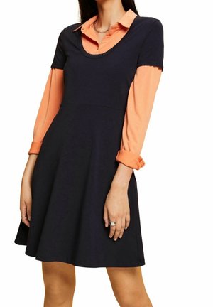 Marineblauw kleed met korte mouwen over een oranje blouse, met een ronde halslijn en een A-lijn silhouet. Glad materiaal en gevouwen manchetten.