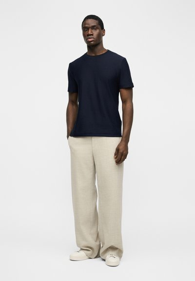 Άνδρας που στέκεται με τα χέρια στις τσέπες, φορώντας μπλε κοντομάνικο πουκάμισο, μπεζ παντελόνι με φαρδιά μπατζάκια και λευκά αθλητικά παπούτσια σε απλό φόντο.