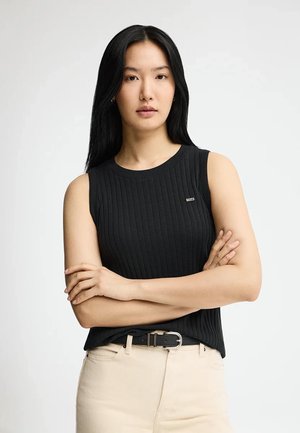 Jeune femme aux longs cheveux noirs, debout les bras croisés, portant un haut noir sans manches à côtes et un pantalon beige avec une ceinture noire.