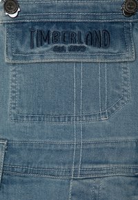 Tessuto in denim blu con dettagli cuciti. Presenta una tasca a toppa e la scritta "TIMBERLAND EST. 1973" ricamata sopra. Inclusi due bottoni in metallo.