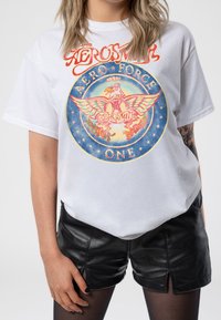 White cotton t-shirt featuring a colorful circular graphic with an eagle, text "Aerosmith," and stars. Paired with black leather shorts.