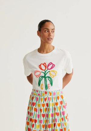 Mujer con una camiseta blanca con bordado de tulipanes de colores y una falda a juego cubierta con un patrón multicolor de tulipanes, de pie contra un fondo liso.