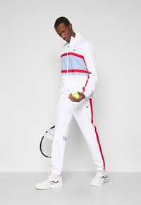 Λευκή φόρμα με κόκκινες και ανοιχτό μπλε οριζόντιες ρίγες, που διαθέτει κολάρο με φερμουάρ και πλάγιες λεπτομέρειες. Το μοντέλο κρατάει μια μπάλα τένις και μια ρακέτα.