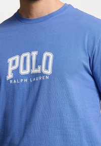 Světle modré bavlněné tričko s výrazným bílým nápisem "POLO" a "RALPH LAUREN" pod ním. Kolo výstřih, krátké rukávy, hladký povrch.