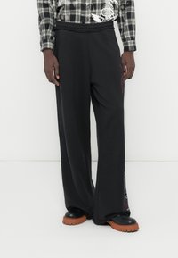 Μαύρο παντελόνι φόρμας με φαρδιά γραμμή, κατασκευασμένο από μαλακό ύφασμα, με ελαστική μέση, πλαϊνές τσέπες και κόκκινες γραφικές λεπτομέρειες.