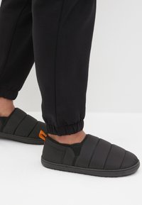 Chaussons noirs rembourrés avec une finition lisse, dotés d'un design à enfiler et d'une languette orange au talon. Portés avec un pantalon de jogging noir.