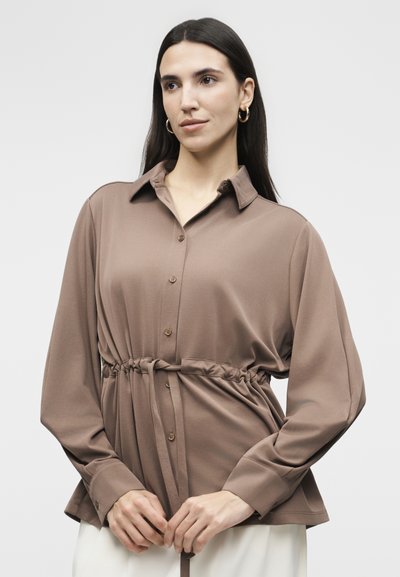 Γυναίκα με μακριά σκούρα μαλλιά, φορώντας χρυσούς κρίκους στα αυτιά και ένα χαλαρό καφέ πουκάμισο με κουμπιά και σκοινί στη μέση, που στέκεται μπροστά σε απλό φόντο.
