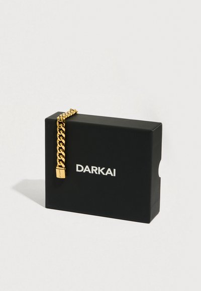 Zlatý řetízkový náramek ležící na černé krabičce. Náramek má robustní design s malým zámkem označeným "DARKAI".