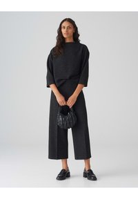 Zwart tekstuur outfit met wijde broekspijpen en een losse trui. Houdt een kleine, doorgestikte zwarte handtas vast. Vlakke zwarte loafers.