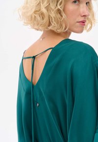 Niebieska jedwabna bluzka z szerokim otwarciem z tyłu, związanymi paskami i dekoracyjnym guzikiem, prezentująca gładką fakturę i elegancki design.