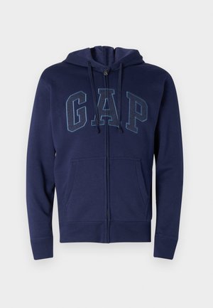Navy blue zip-up hoodie made of cotton blend with front pockets. Features "GAP" logo in lighter blue, adjustable drawstring hood, and ribbed cuffs.