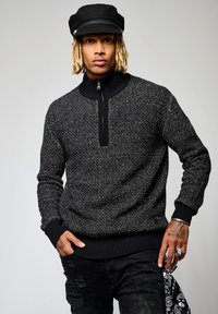 Pull en tricot noir et blanc à motifs, avec un col montant, une demi-fermeture éclair et des poignets côtelés. Porte avec un jean noir déchiré et une casquette noire.