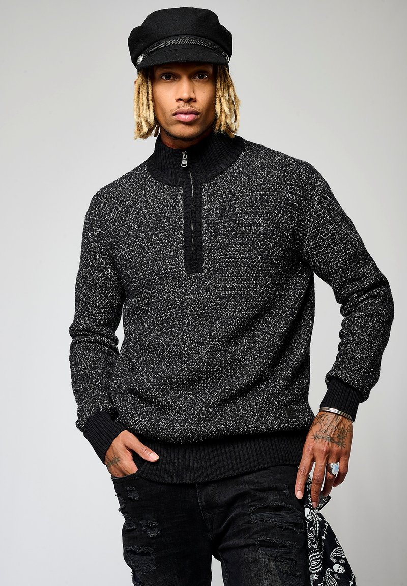 Pull en tricot noir et blanc à motifs, avec un col montant, une demi-fermeture éclair et des poignets côtelés. Porte avec un jean noir déchiré et une casquette noire.