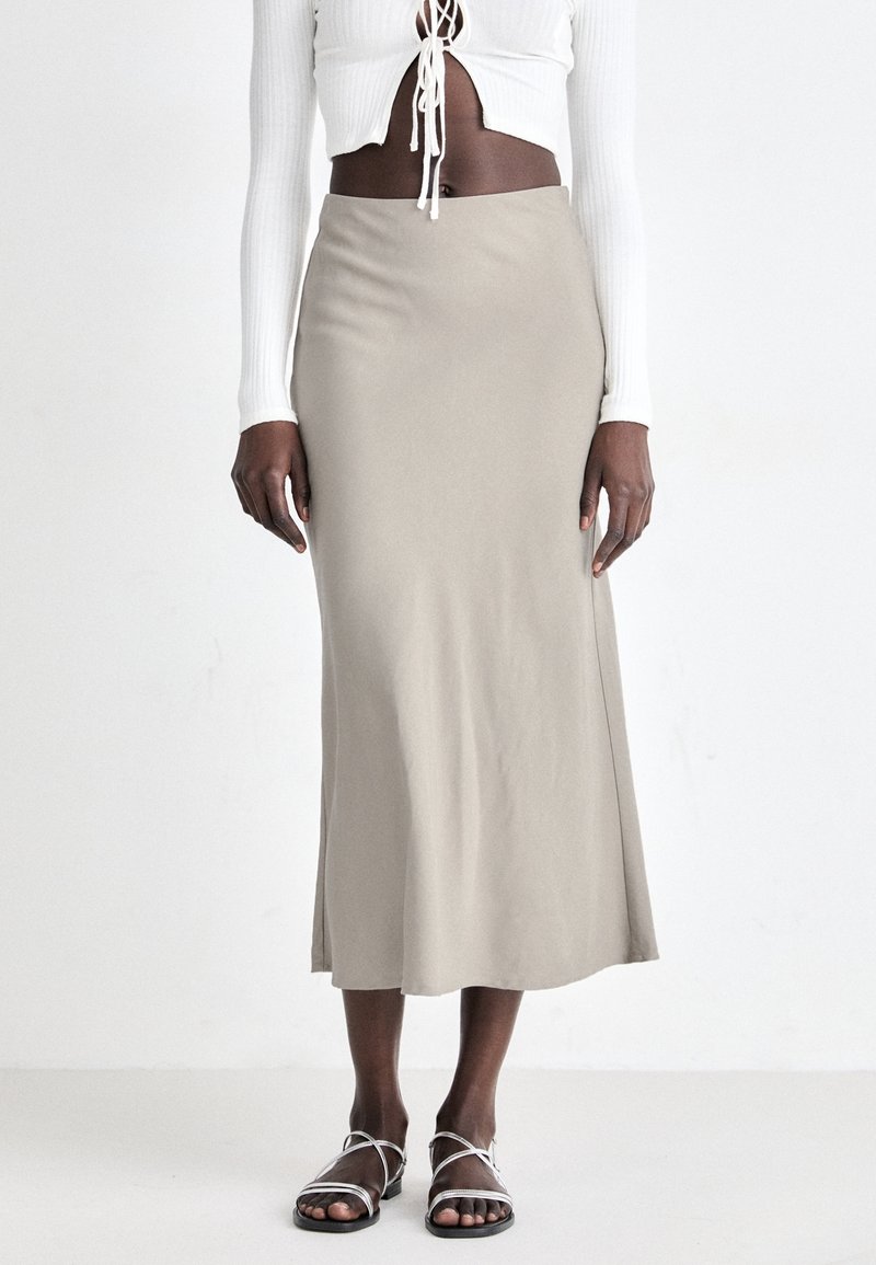 Personne portant une jupe midi beige, un top court blanc à manches longues avec des liens à l'avant et des sandales plates blanches à brides, debout sur un sol blanc.