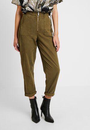 Personne portant un pantalon taille haute vert olive avec poches avant et revers roulés, associé à des bottines noires à talons.