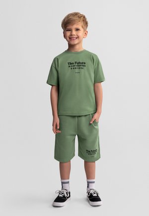 Uśmiechnięty chłopiec o blond włosach, ubrany w zielone spodenki i koszulkę z napisem „Przyszłość dopiero się zaczyna”, czarne trampki i białe skarpetki.