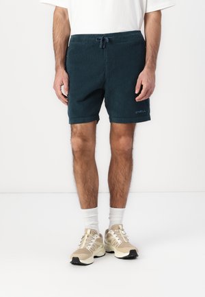 Blue corduroy shorts with an elastic waistband and drawstring, featuring side pockets and a logo detail. Worn with beige sneakers and white socks.