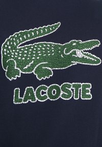 Zelený logo krokodýla s prasklým vzorem a text "LACOSTE" v bílé a zelené barvě na tmavě modrém pozadí. Design má texturovaný vzhled.