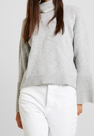 Woman wearing a light grey knit sweater with ribbed cuffs and hem, paired with high-waisted white pants against a plain background.