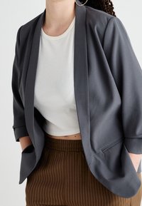 Šedý blazer s přizpůsobeným střihem a širokými klopami, nošený přes bílé příčkové crop top, v kombinaci s hnědými kalhotami s jemným pruhem.