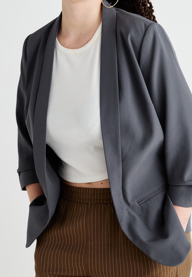 Šedý blazer s přizpůsobeným střihem a širokými klopami, nošený přes bílé příčkové crop top, v kombinaci s hnědými kalhotami s jemným pruhem.