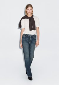 Biała koszula z krótkim rękawem, ciemnobrązowy szalik zawiązany na szyi, niebieskie dżinsy i czarne buty na obcasie, stojąca na białym tle.