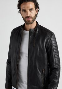 Zwarte leren jas met een ritssluitingen aan de voorkant, gestikte schouderaccenten en zijzakken. Glad oppervlak en een slim fit ontwerp.