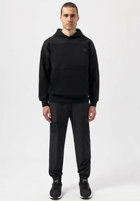 Sudadera negra con detalles de malla y un bolsillo frontal, combinada con pantalones cargo negros con bolsillos laterales. Ambos artículos tienen un ajuste holgado.