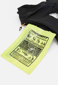 Gilet nero con una tasca con zip che mostra una vivace scheda informativa di emergenza gialla con icone e testo sulla sicurezza in montagna.