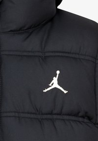 Veste rembourrée noire avec un collier montant et un logo Jumpman blanc sur la partie gauche de la poitrine. Texture lisse avec des détails de couture matelassée.