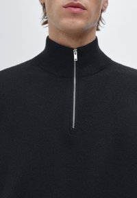 Zwarte zip-up trui met een hoge kraag, gemaakt van gestructureerd breiwerk. Heeft een zilveren ritsdetail aan de voorkant.