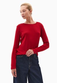 Czerwona koszulka z długim rękawem i okrągłym dekoltem, z płaskimi szwami, w połączeniu z ciemnoniebieskimi dżinsami o wysokim stanie. Gładka tekstura, dopasowany krój.