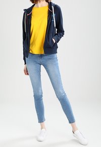 Navy zip-up hoodie with white drawstrings, yellow crew neck t-shirt, light blue skinny jeans, and white sneakers. Simple casual style.