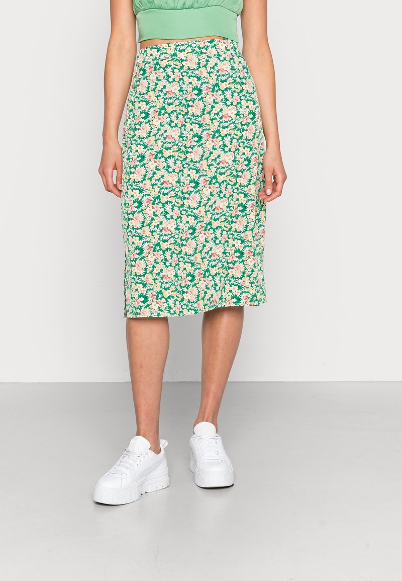 Femme portant une jupe verte fleurie longueur genou, un crop top vert et des baskets blanches à plateforme, debout sur un sol gris avec un fond blanc.