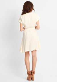 Vestido de crema de manga corta con cintura ajustada y falda acampanada. La parte trasera presenta una costura sutil y un cinturón de tela. Complementado con botas de tacón en color marrón.
