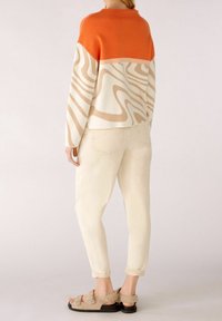 Maglione lavorato a maglia con sezione superiore arancione e motivo ondulato beige, abbinato a pantaloni beige chiaro e sandali morbidi color tan con fibbia.