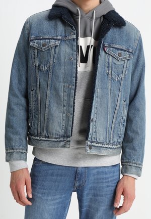 Mand iført blå denimjakke med sort lammeskindskrave over grå hættetrøje og blå jeans, stående mod en ensfarvet hvid baggrund.