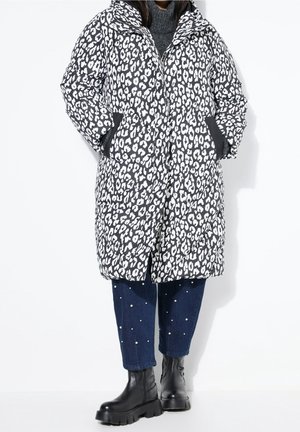Personne portant un manteau matelassé imprimé léopard noir et blanc, un jean court bleu avec des perles et des bottines noires épaisses, debout sur fond blanc.