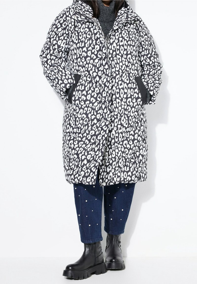 Personne portant un manteau matelassé imprimé léopard noir et blanc, un jean court bleu avec des perles et des bottines noires épaisses, debout sur fond blanc.