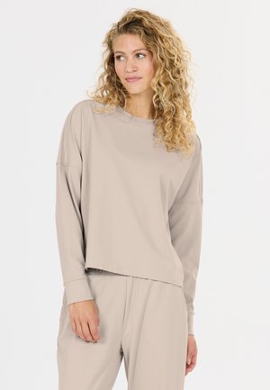Kvinde med krøllet blondt hår iført løs beige langærmet top og matchende bukser, der ser en smule mod hendes venstre side.