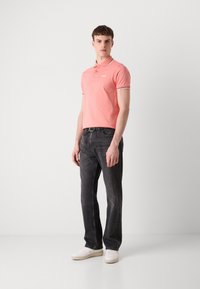 Polo shirt rose clair avec logo blanc, associé à un jean gris foncé et des baskets blanches. Présente un col classique et des accents rayés sur les manches.