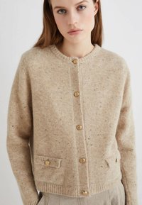 Cardigan in lana beige con scollo tondo, bottoni dorati e due tasche frontali. Texture morbida con leggere macchie throughout.