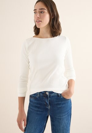 Witte geribbelde lange mouw top met een bateauhals, gedragen met blauwe denim jeans met een voorzak en een lichte vervaging.