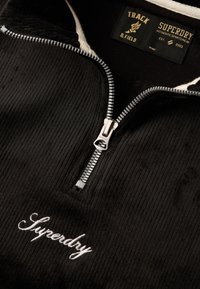 Pullover in velluto a coste nero con zipper silver, dotato di colletto in pelliccia sintetica e logo ricamato "Superdry". L'etichetta indica "TRACK & FIELD".