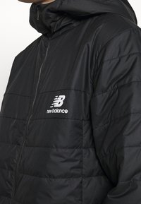 Svart vadderad jacka med dragkedjestängning, hög krage och New Balance-logotyp på vänster bröst. Har en slät, glänsande textur.