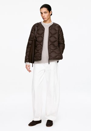 Vrouw die een bruin gewatteerd jack draagt over een beige trui, witte wijde broek, donkere schoenen, en een bruine schoudertas bij zich heeft.