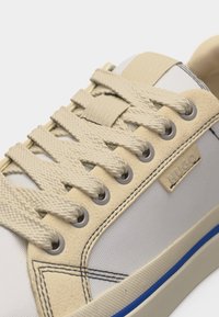 Μπεζ και λευκό αθλητικό παπούτσι με υφασμένα κορδόνια, μεταλλικά ρουλεμάν, ραμμένες λεπτομέρειες και μικρή ετικέτα με το λογότυπο "HUGO" στην πλευρά.