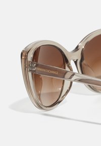 Cat-eye solbriller med gradient brune linser og gjennomsiktig brun ramme. Gull "Armani Exchange" merkevare på den gjennomsiktige stang.