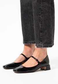 Schwarze Lackleder Schuhe mit einem niedrigen Absatz, spitzer Zehenform und Knöchelriemen mit einer Schnallenauffassung. Getragen mit dunklen Jeans.