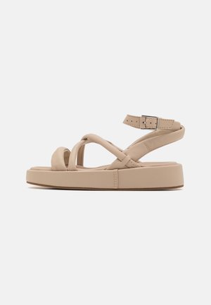Sandali beige con suola spessa, caratterizzati da un design a strap incrociate e una cinghia alla caviglia regolabile. Texture liscia e forma arrotondata.